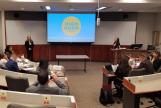 A young woman stands at the front of a lecture-style classroom presenting to those seated around her.